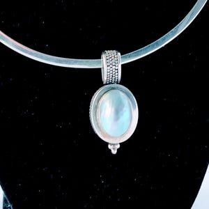 May include: A silver-toned necklace with a pendant featuring a large, oval, iridescent stone. The necklace has a simple, curved design, and the pendant is suspended from a textured bail. The necklace is displayed on a black jewelry bust.