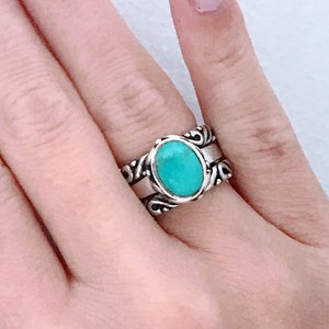 May include: A silver ring with a turquoise oval stone set in a decorative band. The ring is worn on the middle finger of a hand with light pink nail polish.