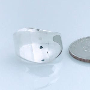 May include: A shiny, silver ring with a wide band and a smooth, polished surface. The ring is next to a quarter coin for size comparison. The ring's design is simple and modern, reflecting light.