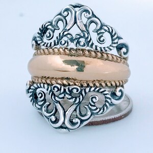 May include: A silver ring with a gold band and intricate filigree design. The ring is set on a white background.