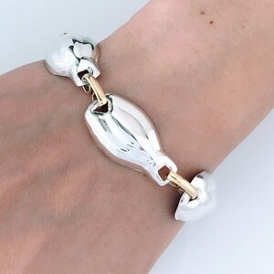 May include: A silver and gold-toned bracelet with a unique link design. The bracelet features large, rounded silver links connected by smaller gold-toned bars. The jewelry is displayed on a person's wrist.