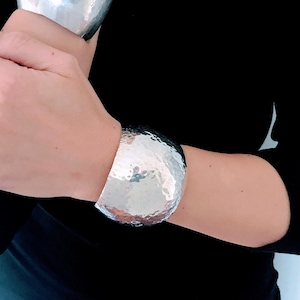 May include: Two silver hammered metal bracelets on a person's wrist. The bracelets are both wide and have a rounded shape.