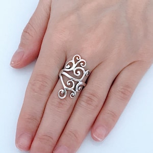 May include: A silver ring with a delicate, swirling design. The ring is worn on the middle finger of a hand.