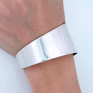 May include: A wide, silver-colored cuff bracelet. The bracelet is a solid band of polished metal, reflecting light. The cuff is worn on a wrist, showcasing its minimalist design and smooth, reflective surface. A modern piece of jewelry.