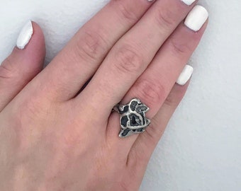 May include: A silver ring with a detailed design featuring a heart and other abstract shapes. The ring is worn on a hand with white nail polish.