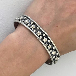 May include: A silver bangle bracelet with a textured design featuring a repeating pattern of small circles and stars.