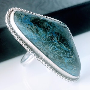 May include: A silver ring featuring a large, elongated, diamond-shaped stone with a deep blue and black marbled pattern. The stone is set in a smooth silver bezel surrounded by a decorative beaded silver wire border on a simple silver band.
