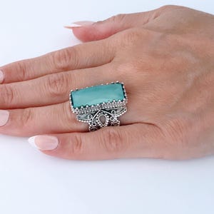 May include: A silver ring with an aquamarine-colored rectangular stone. The ring has an ornate, filigree design around the stone and on the band. The ring is on a person's hand with manicured nails.