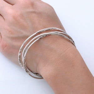 May include: A set of three silver bangles with a hammered texture. The bangles are stacked on a person's wrist.