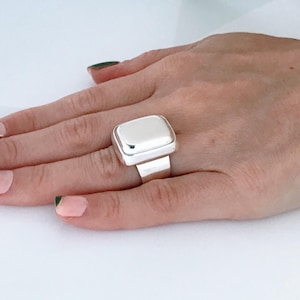 May include: A silver ring with a large, rectangular, flat top. The ring is on a person's finger.