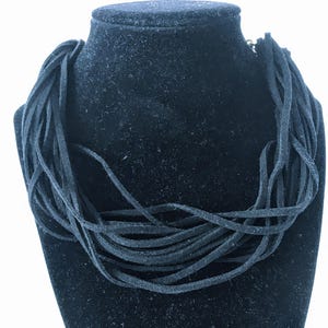 May include: A multi-strand necklace composed of numerous thin, black suede-like cords. The cords are arranged in a layered, overlapping fashion, creating a textured, circular collar shape that sits around the neck area with a matte finish.
