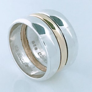 May include: A wide band ring featuring a polished silver exterior and a smooth gold-toned center band. The interior of the ring is stamped with the text MEXICO 925, indicating the metal composition and origin of this two-tone jewelry piece.