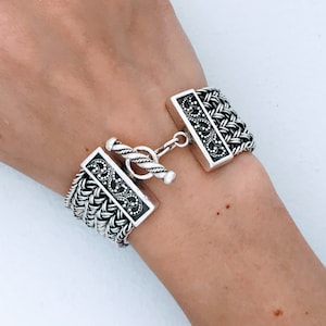 May include: A silver bracelet with a detailed, intricate design. The bracelet has a toggle clasp.