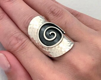 May include: A silver ring with a hammered finish and a black spiral design. The ring is wide and sits high on the finger.