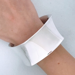 May include: A silver bangle bracelet with a wide, flat, and slightly curved design.