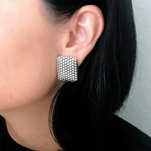 May include: Silver-toned square stud earring with a textured surface. The earring is made up of many small, round, silver-colored beads. The earring is worn on the ear of a person with black hair.