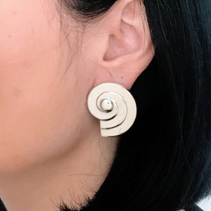 May include: Silver spiral earrings with a rounded center. The earrings are a modern design and are a statement piece.