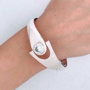 May include: A silver bracelet with a large, clear crystal set in the center. The bracelet is open and has a curved design.