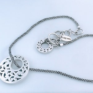 May include: A silver necklace with a twisted chain and a large, circular pendant with a cut-out design. The pendant is attached to the chain with a smaller, circular charm. The necklace has a tag that reads "LOIS HILL".