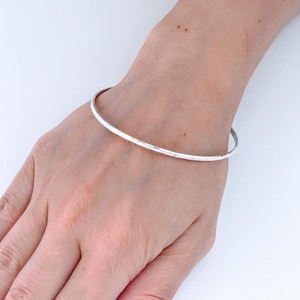May include: A simple, silver-colored bangle bracelet. The bracelet is a thin, smooth band, worn on a person's wrist. The background is a plain white surface. The bracelet is a classic piece of jewelry.