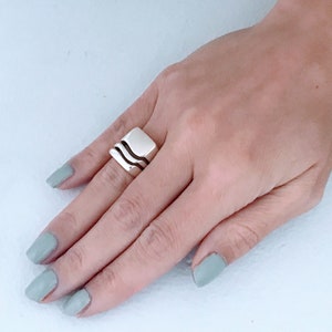 May include: A square-shaped silver ring with two dark bands. The ring is on a hand with light green nail polish.