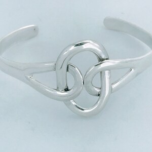 May include: A silver bracelet with a knot design. The knot is a circle with a smaller circle inside it, and the ends of the knot are connected to the bracelet.