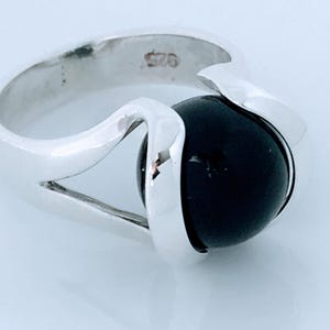 May include: A sterling silver ring with a large, round, black stone. The ring has a modern design with a curved band that wraps around the stone. The ring is marked with "925".