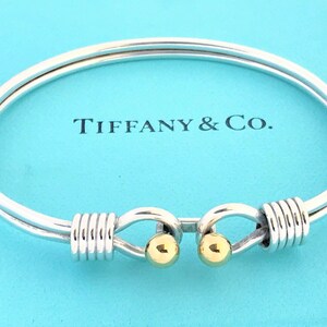 May include: A silver bangle bracelet with two gold accent balls and a twisted design. The bracelet is on a blue background with the text "Tiffany & Co."
