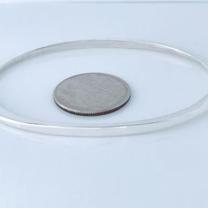 May include: A silver-colored bangle bracelet with a circular design. A quarter coin is placed inside the bracelet. The bracelet is made of a shiny, reflective metal. The background is a plain white surface.