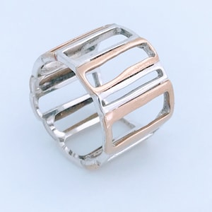 May include: A silver and rose gold ring with a geometric design. The ring features a series of rectangular openings, with alternating silver and rose gold bands. The ring is a wide band style.