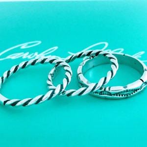 May include: Three silver rings with a twisted rope design. The rings are stacked on top of each other on a turquoise background.
