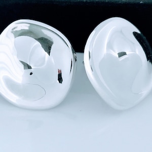 May include: A pair of polished, silver-colored stud earrings. Each earring has a smooth, rounded, organic shape, reflecting light. The earrings are displayed against a white surface with a dark background.
