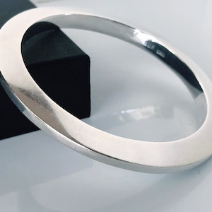 May include: A silver-colored, square-edged bangle bracelet. The bracelet has a smooth, polished surface and a minimalist design. The jewelry is displayed on a white surface with a black box in the background. The bracelet is approximately 2.5 inches in diameter.