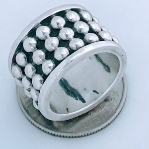 May include: A silver ring with a wide band and a textured design of small, round beads. The ring is shown next to a US quarter for size comparison.