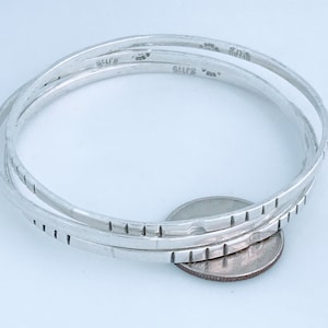 May include: Three silver bangles with a textured design. The bangles are stacked on top of each other and are marked "STERLING 925".