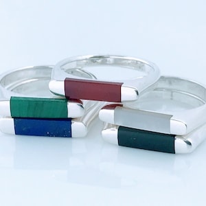 May include: Four silver rings with different colored stone inlays. The rings are stacked on top of each other. The stones are red, green, blue, and gray.