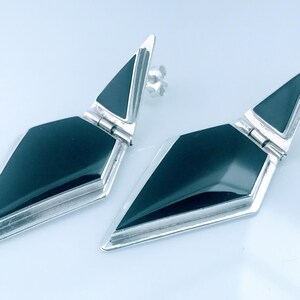 May include: A pair of geometric earrings with a silver-tone metal frame and a black, diamond-shaped center. The earrings have a post-back closure. The design is modern and minimalist.