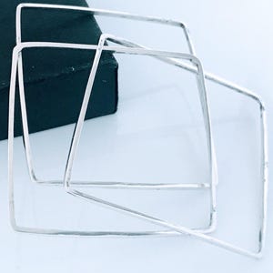May include: Three silver square bangles are displayed, with a dark box in the background. The bangles are made of a shiny metal and have a minimalist design. The jewelry is set against a light background.