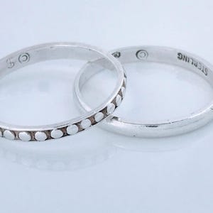 May include: Two silver rings. One ring has a series of small, raised dots around the band. The other ring is smooth and has the word "STERLING" stamped on it. Both rings are on a white surface.