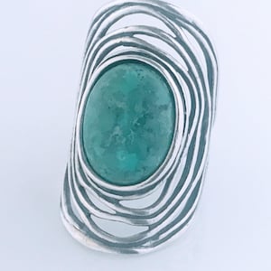 May include: A silver ring featuring a large, oval-shaped, mottled green stone set in the center. The band is constructed from multiple overlapping, curved silver wires that create an open, cage-like structure surrounding the central gemstone.