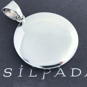 May include: A polished, circular silver pendant with a smooth, reflective surface and a sturdy bail at the top. The pendant is positioned directly above the white text "SILPADA" which features a stylized arrow symbol to the left of the brand name.