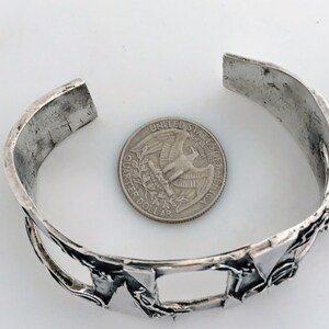 May include: A silver bracelet with a geometric design. The bracelet is open and has a gap in the design. A US quarter is shown for scale.