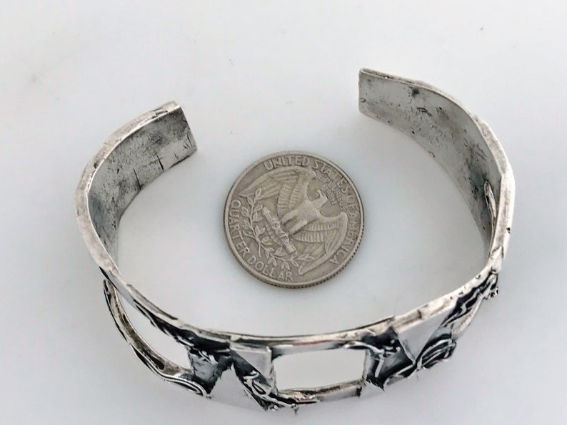 May include: A silver bracelet with a geometric design. The bracelet is open and has a gap in the design. A US quarter is shown for scale.