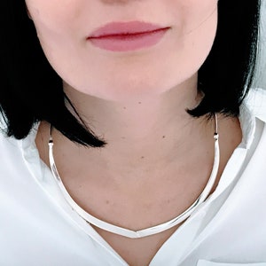 May include: A silver choker necklace with a curved, V-shaped design. The necklace is worn by a person with black hair.