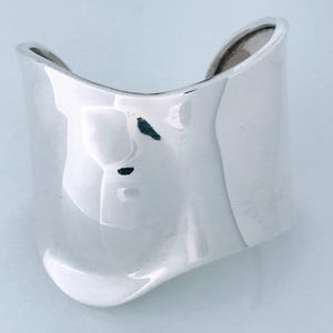 May include: A wide, polished silver cuff bracelet with a smooth, undulating surface. The metal has a reflective, mirror-like finish with gentle curves that create a wave-like silhouette. The bracelet features an open back design for easy wear.