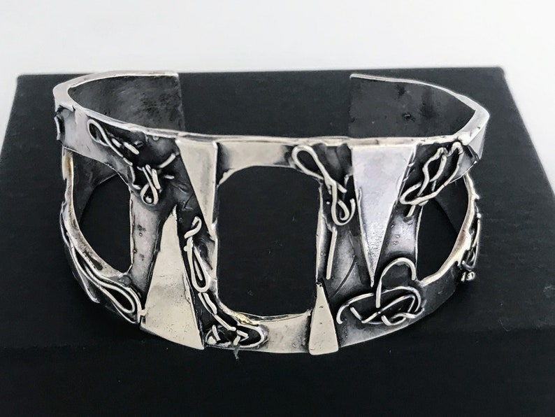 May include: A silver cuff bracelet with a geometric design. The bracelet has a textured surface and is made of metal.