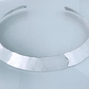 May include: A silver-colored collar necklace with a hammered texture. The open-ended choker has a wide, flat band that tapers towards the ends. The jewelry piece is displayed on a light-colored surface.