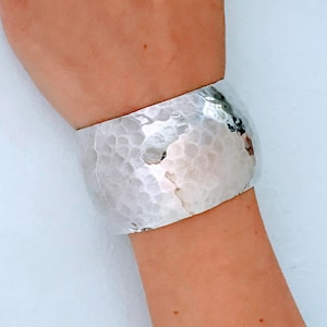 May include: A silver hammered cuff bracelet. The bracelet is wide and has a rounded shape. It is made of a smooth, shiny metal.