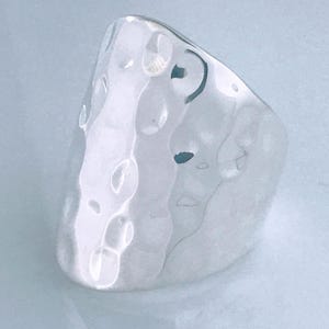 May include: A wide, polished silver ring featuring a distinct hammered texture across its entire outer surface. The metal has a reflective, high-shine finish with multiple shallow indentations that create a varied, light-catching appearance.