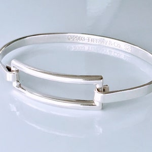 May include: A silver Tiffany & Co. bangle bracelet with a rectangular opening. The bracelet is engraved with the text "©2003 TIFFANY & CO. 925".